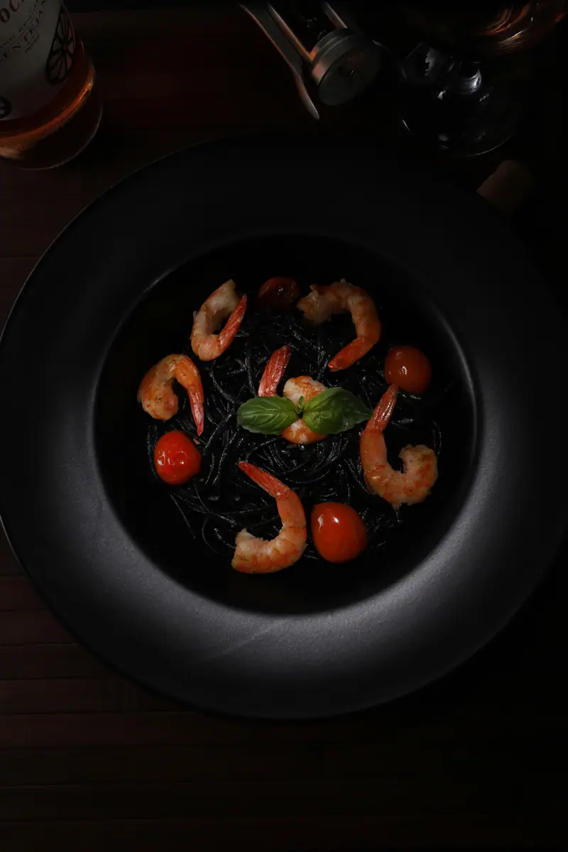 Camarões, massa preta e tomate-cereja em prato preto, vistos de cima