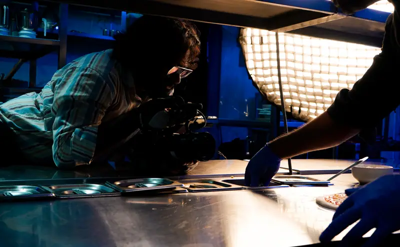 Fotógrafo enquadrando na bancada de inox de uma cozinha profissional, com softbox de grade ao lado